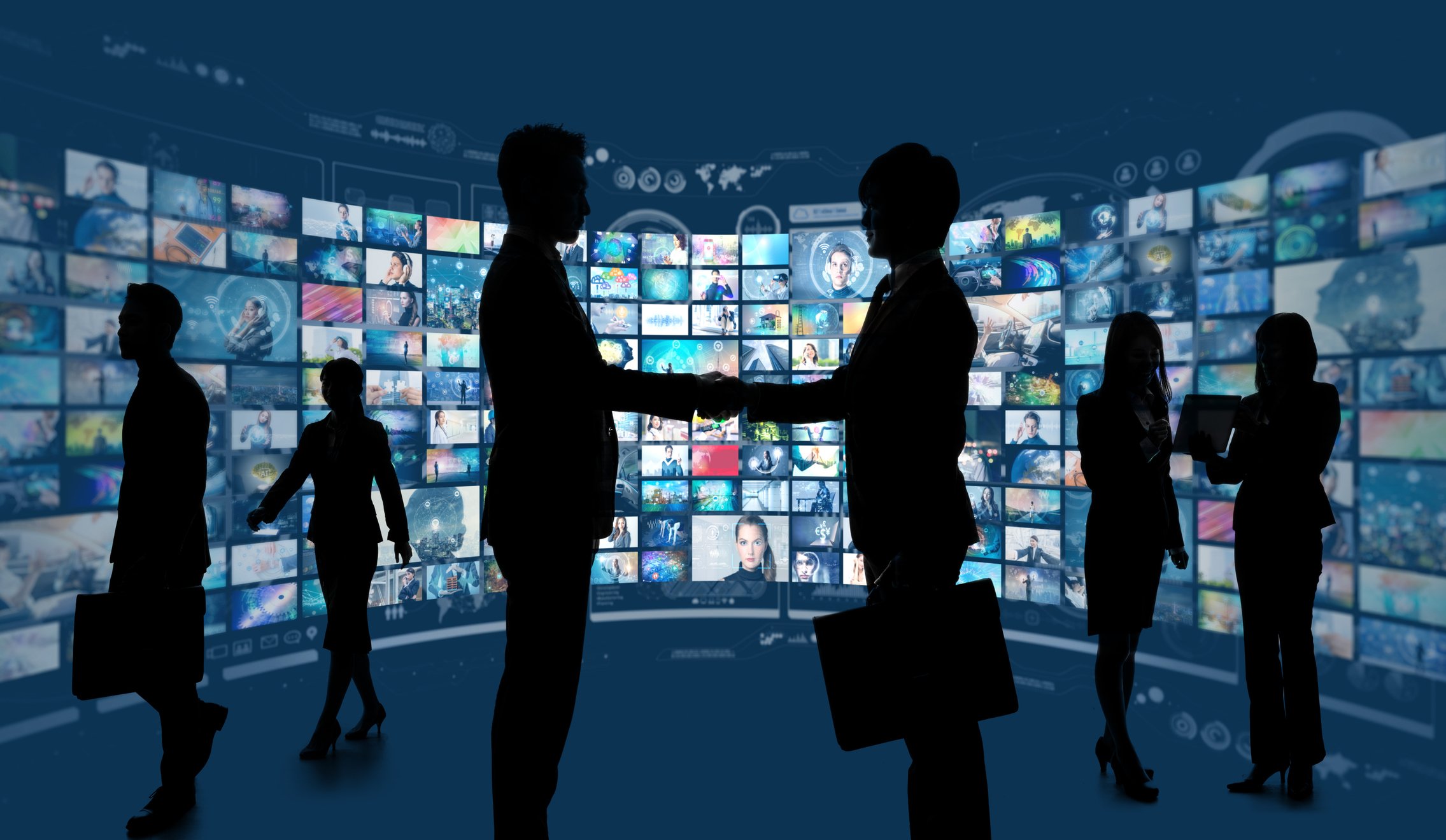 Two businessmen shaking hands in silhouette in fornt of dozens of TV screens.