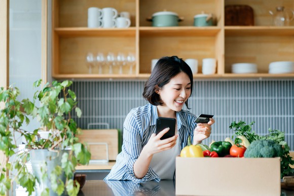 A young woman in a kitchen makes a purchase on her mobile phone.