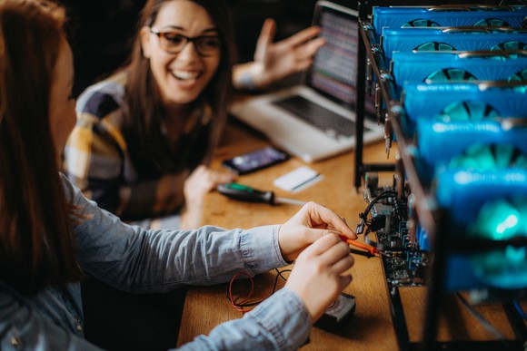 Two young women work on a crypto mining rig while laughing.