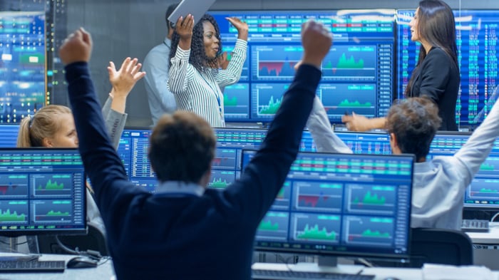 stock traders cheering in room with many monitors showing stock charts.