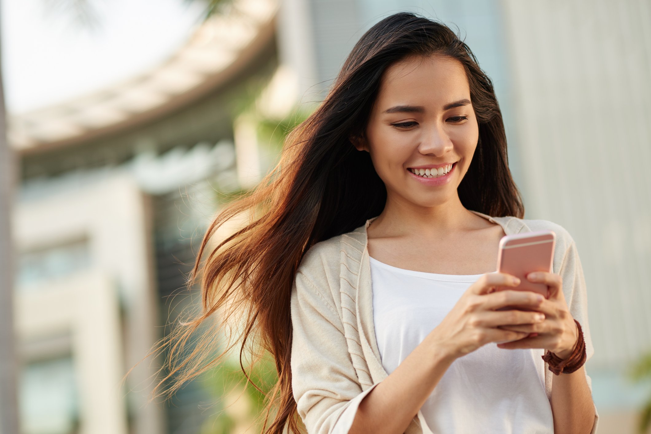 A smiling person walking outdoors texts on their mobile phone.