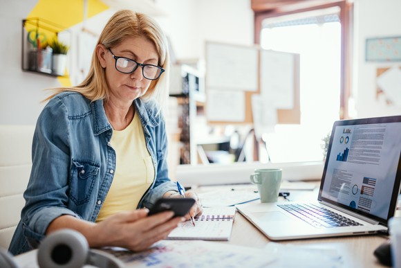 A woman looking at her phone while sitting in front of a laptop.