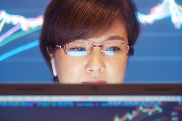 Person wearing glasses in front of a PC screen with a large stock chart displayed in the background.