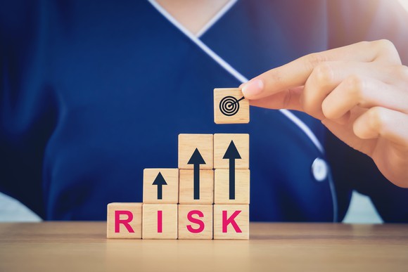 A person placing a wood block showing an arrow hitting a bulls-eye on top of wood blocks with arrows pointing up with other blocks spelling the word "Risk." 