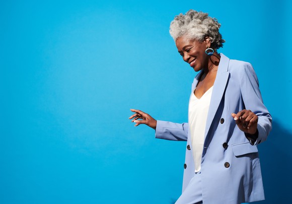 A smiling older woman  dancing for joy against a blue background.