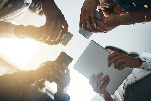 Four people stand in circle and look at their electronic devices seen from ground up. 