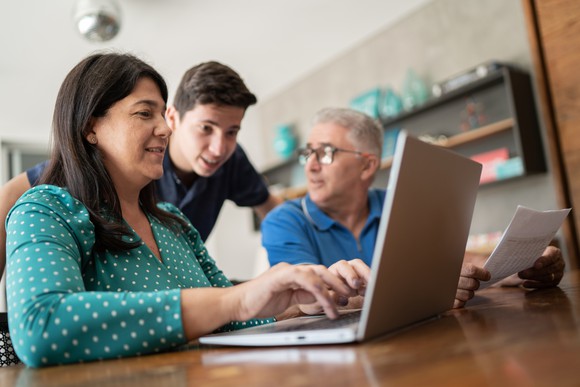 Young person leans over two adults' shoulder while they look at a laptop computer screen.