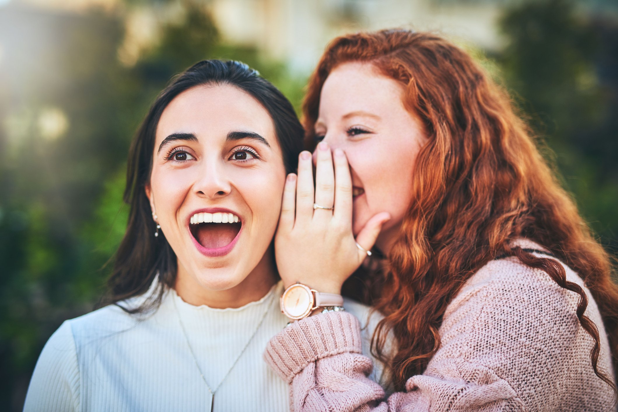One woman whispering into the ear of another woman, who is pleasantly surprised by what she is hearing. 