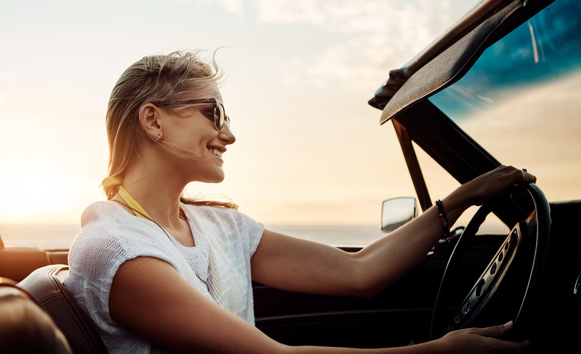 Smiling woman driving convertible as the sun sets in the distance.