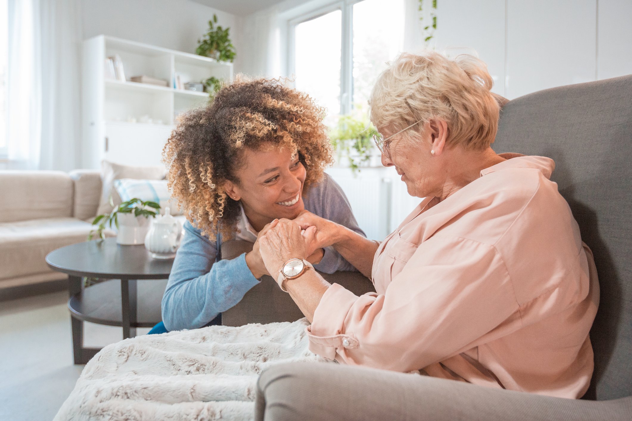 A smiling caregiver comforts an elderly woman sitting in a chair.