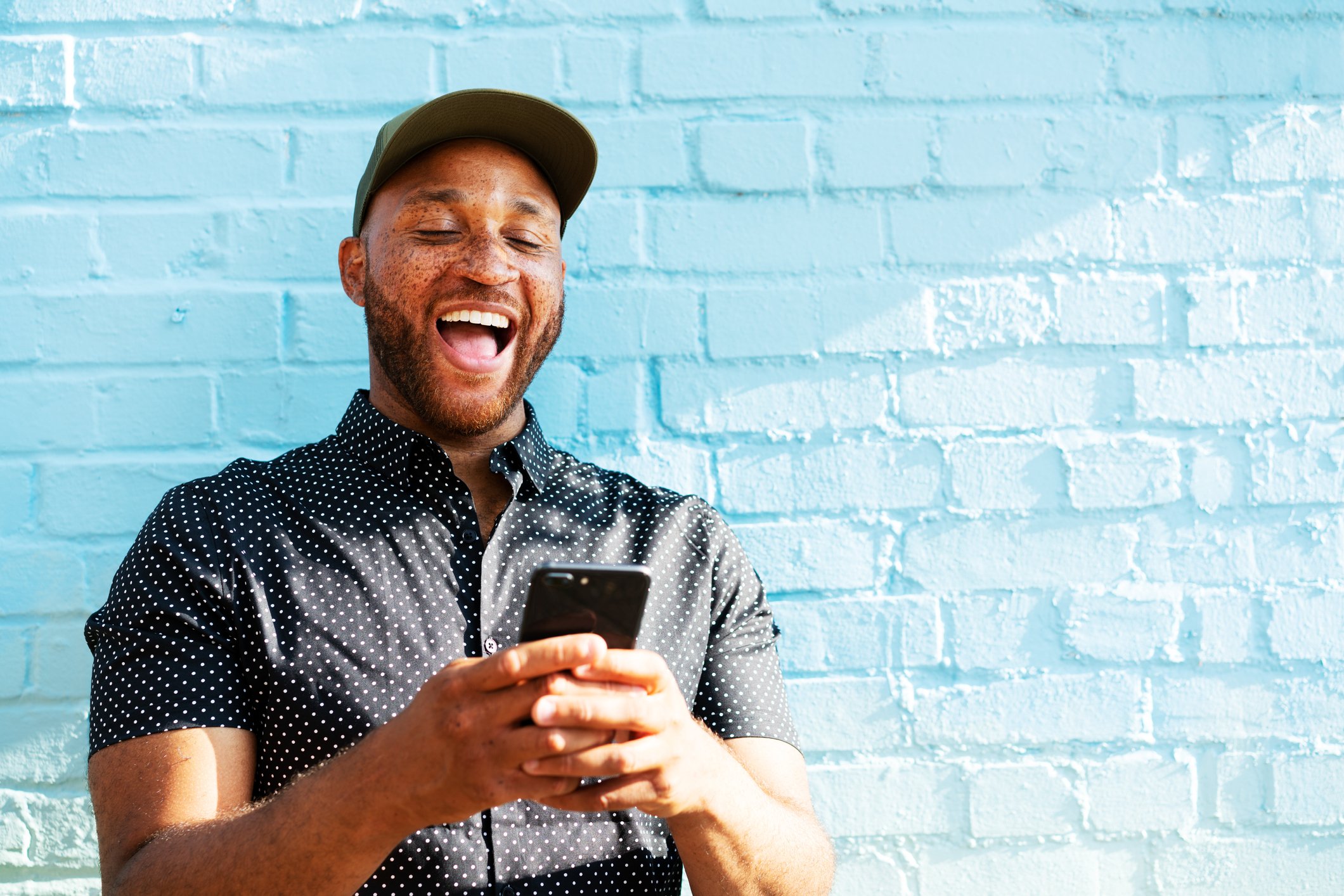 A person appears to laugh out loud while looking at a smartphone and standing in front of a blue brick wall.