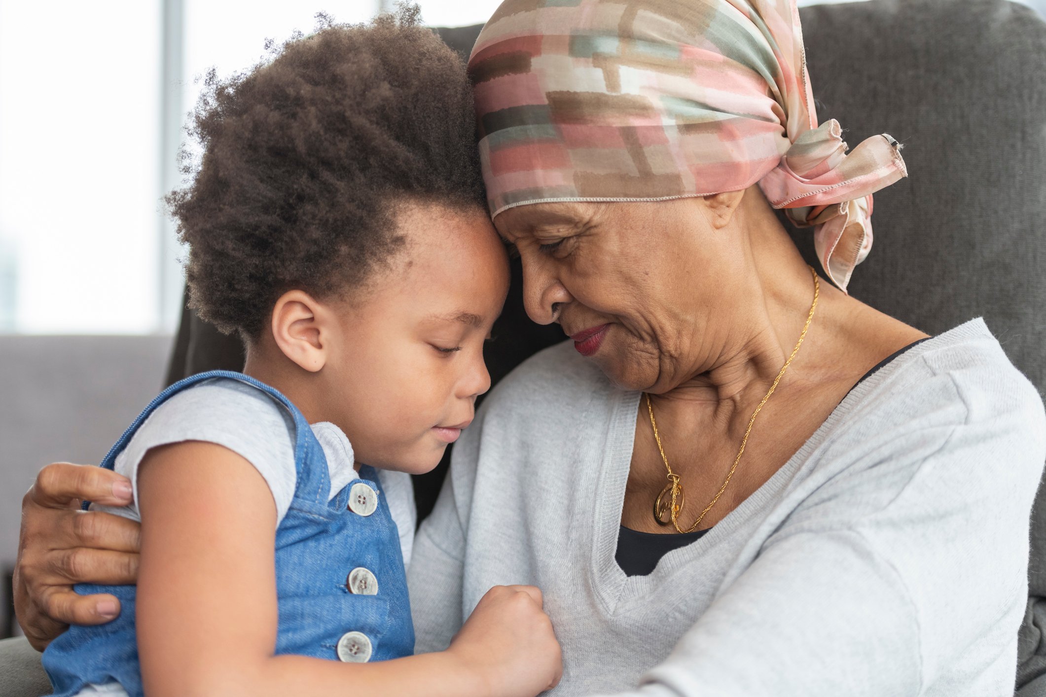 An elderly cancer patient and a young child holding each other in a chair with their foreheads touching.