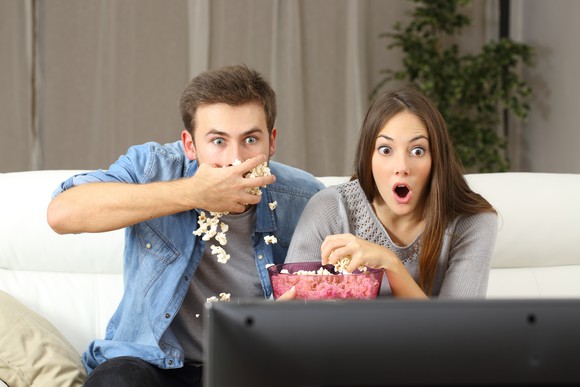 A young couple eating popcorn and looking shocked at the TV screen.
