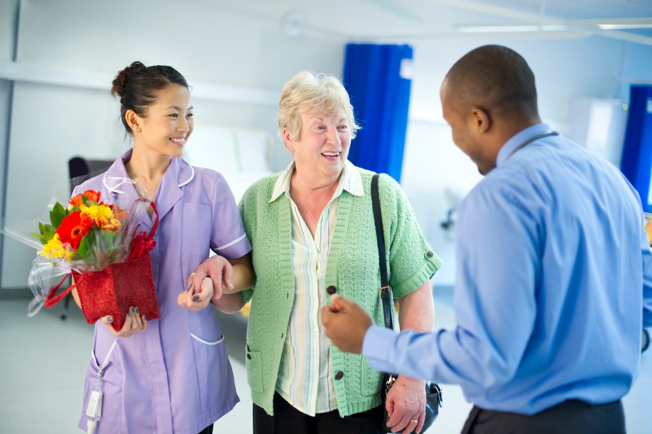 A patient, a nurse holding flowers, and a doctor talking as the patient leaves the hospital unit.