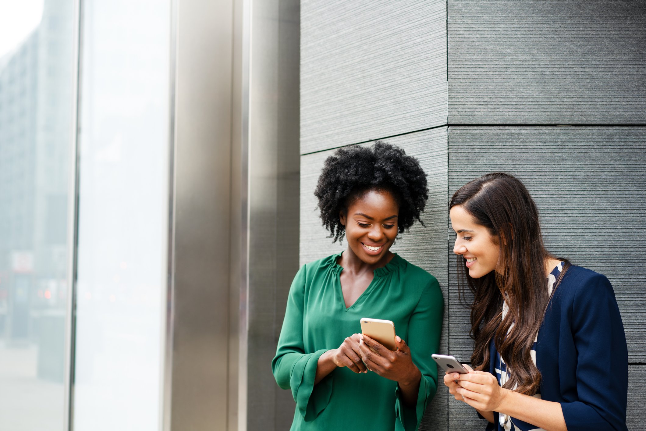 Two friends looking at social media posts on each other's phones as they stand outside of a building. 