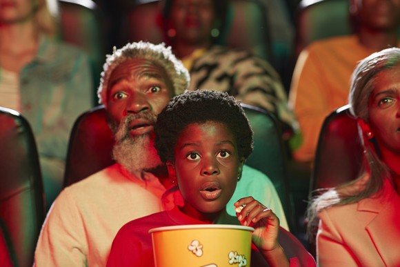 A grandfather and young grandchild on his lap watch a movie in a theater. 