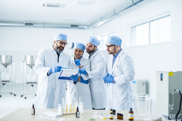 Four chemists examine a clipboard with data while standing in their laboratory.