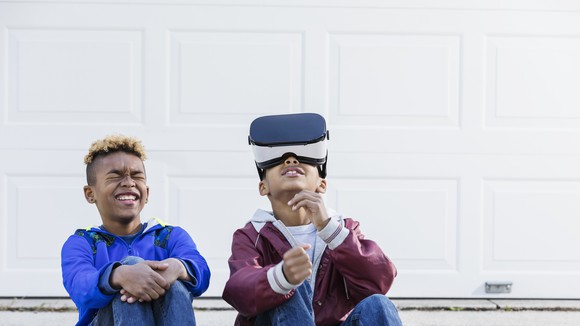 Two kids sitting together, with one wearing a virtual reality headset.