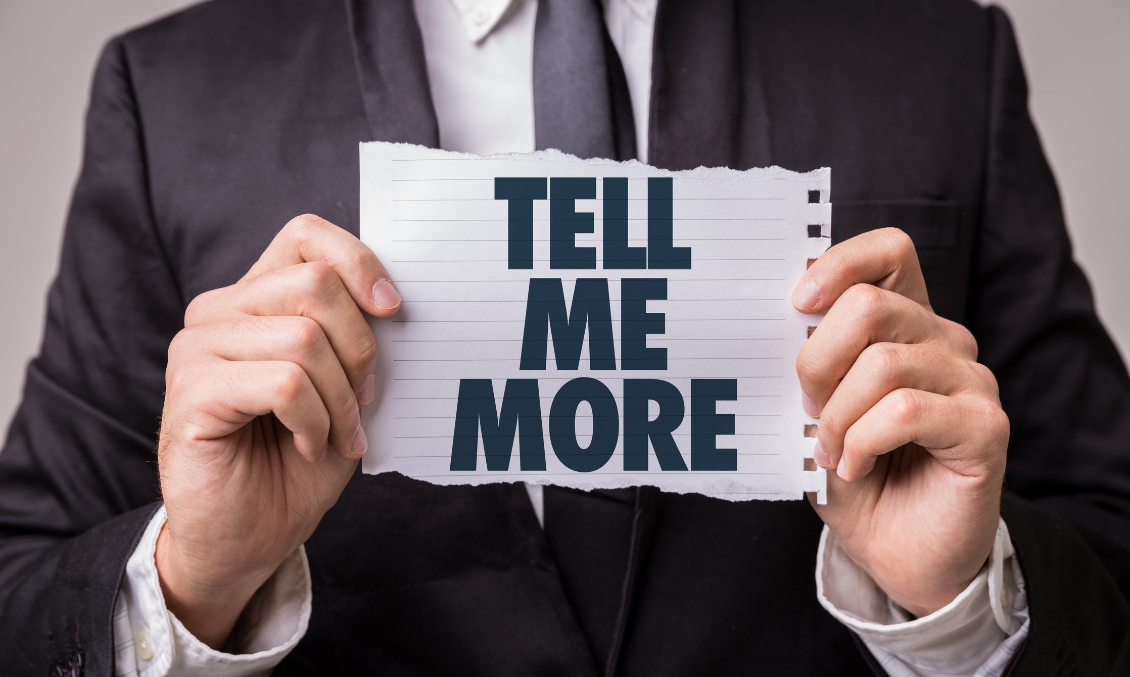 Someone in a suit is holding up a sign that says tell me more.