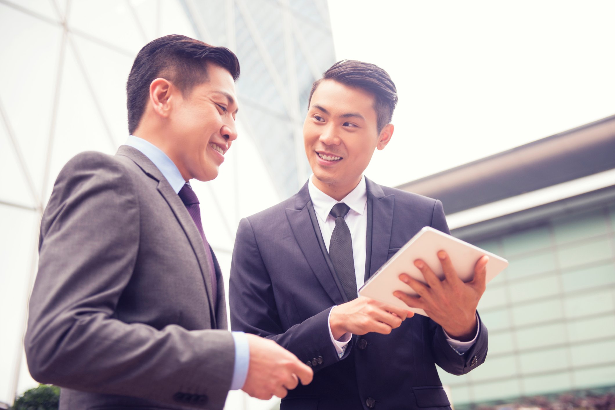 Two young men in business suits converse looking at a tablet. 