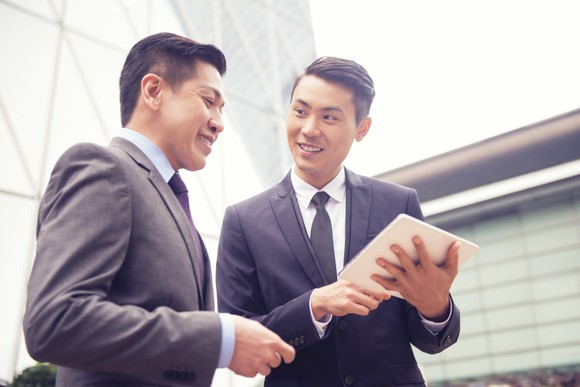 Two young men in business suits converse looking at a tablet. 