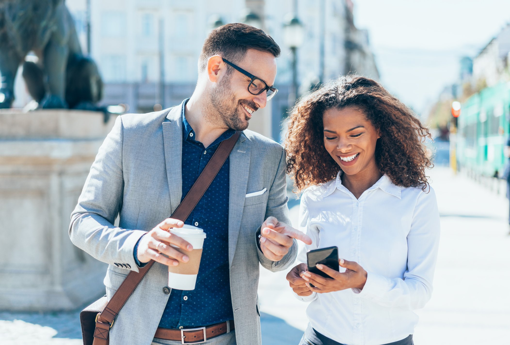 Two people laughing at something on a smartphone while walking down the street.