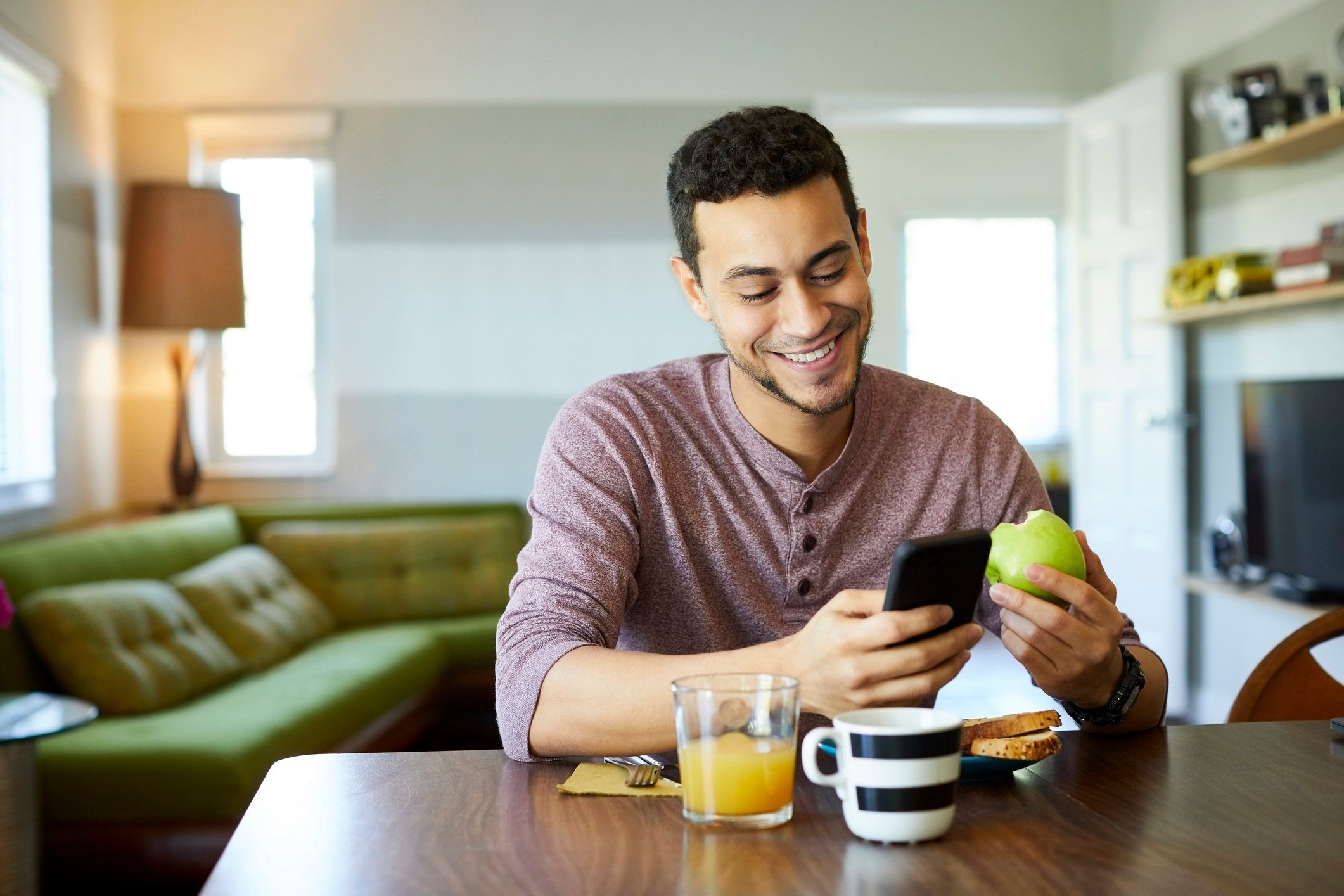 A person sitting at a table eating an apple while looking at his smartphone.