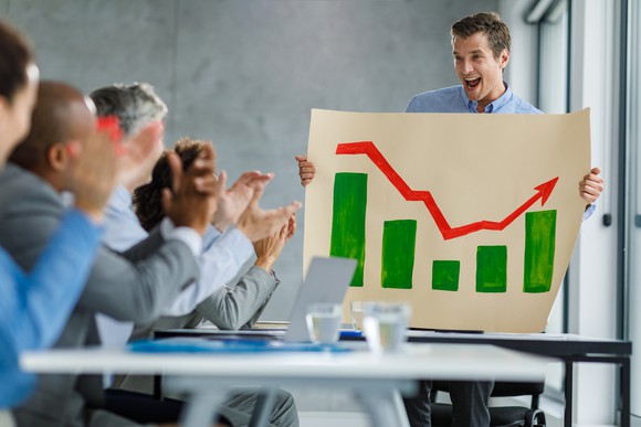 A man holding a chart with a red stock arrow going up in front of a group of people.