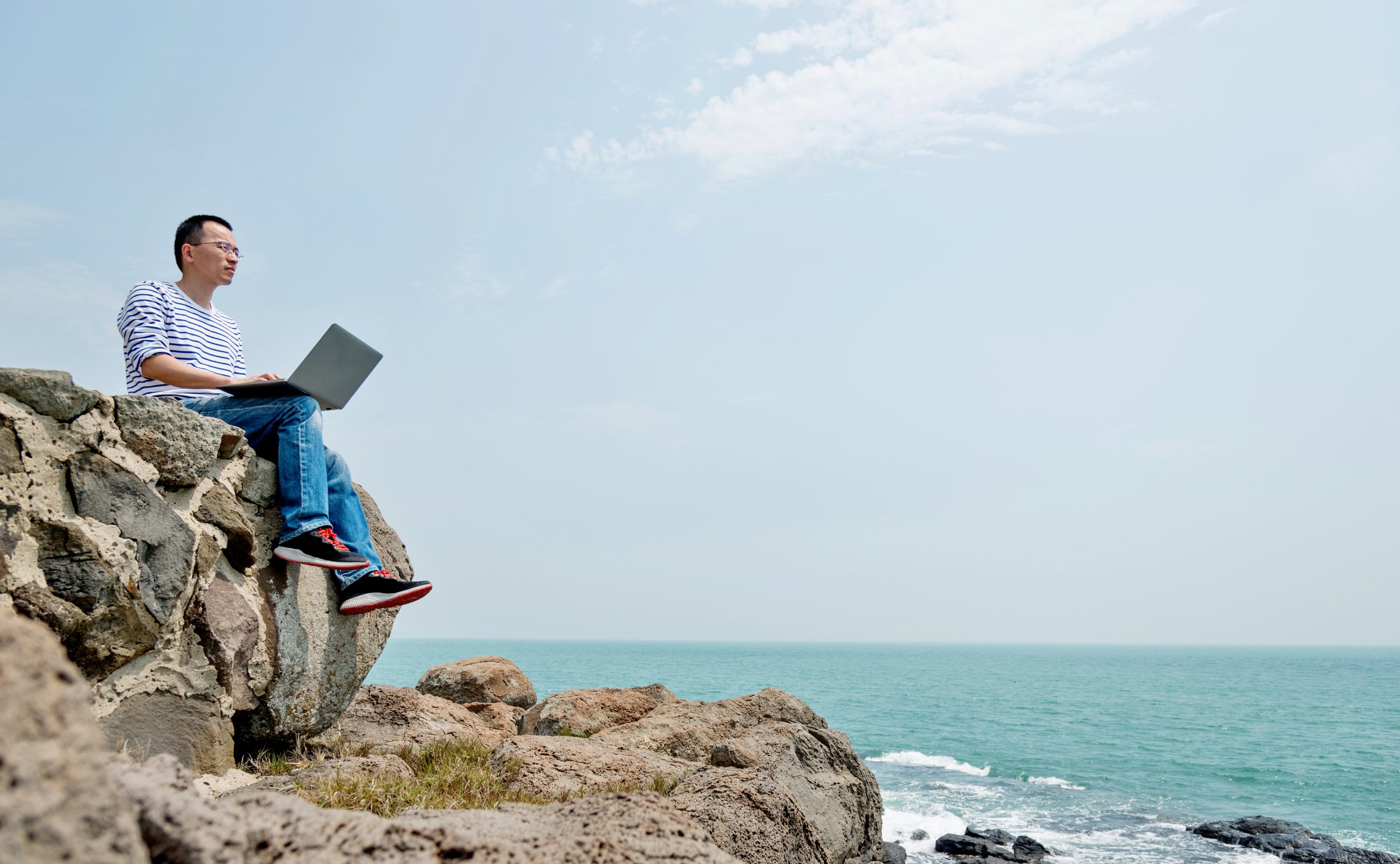 A person uses a laptop while siting on a rocky hilltop overlooking the ocean.