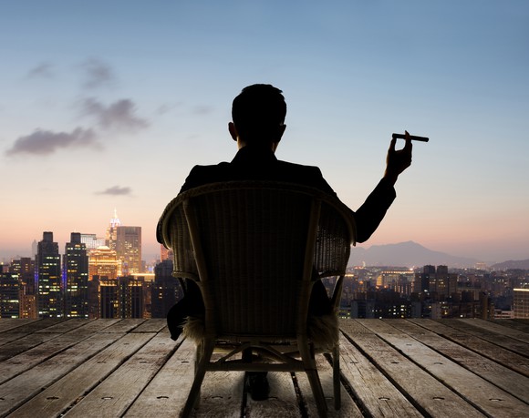 Person sitting in chair on deck overlooking city, holding cigar