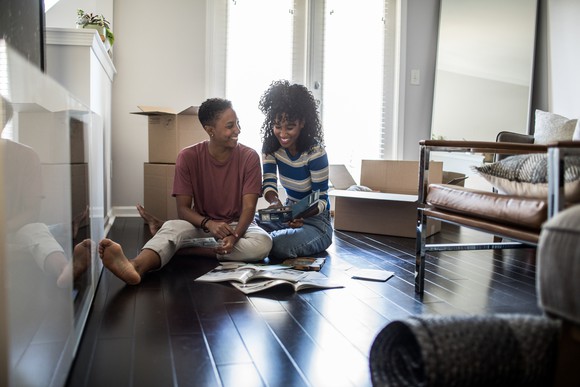 Two people sitting on living room floor smiling and looking at magazines.