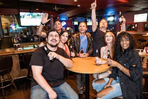 a group of people cheer while sitting around a table holding drinks in a sports bar