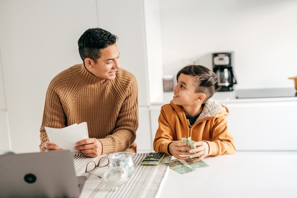 An adult holding papers and a child holding money sitting at table.
