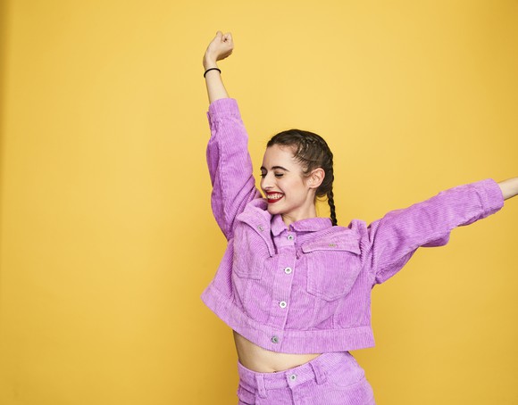 A happy young person wearing a purple outfit dancing against a yellow background.