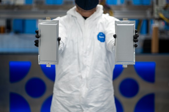 An engineer in a white lab coat holding two rectangular battery cells. 