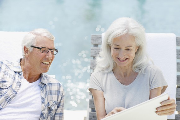 Two people sit by a pool and smile as they look at a laptop.
