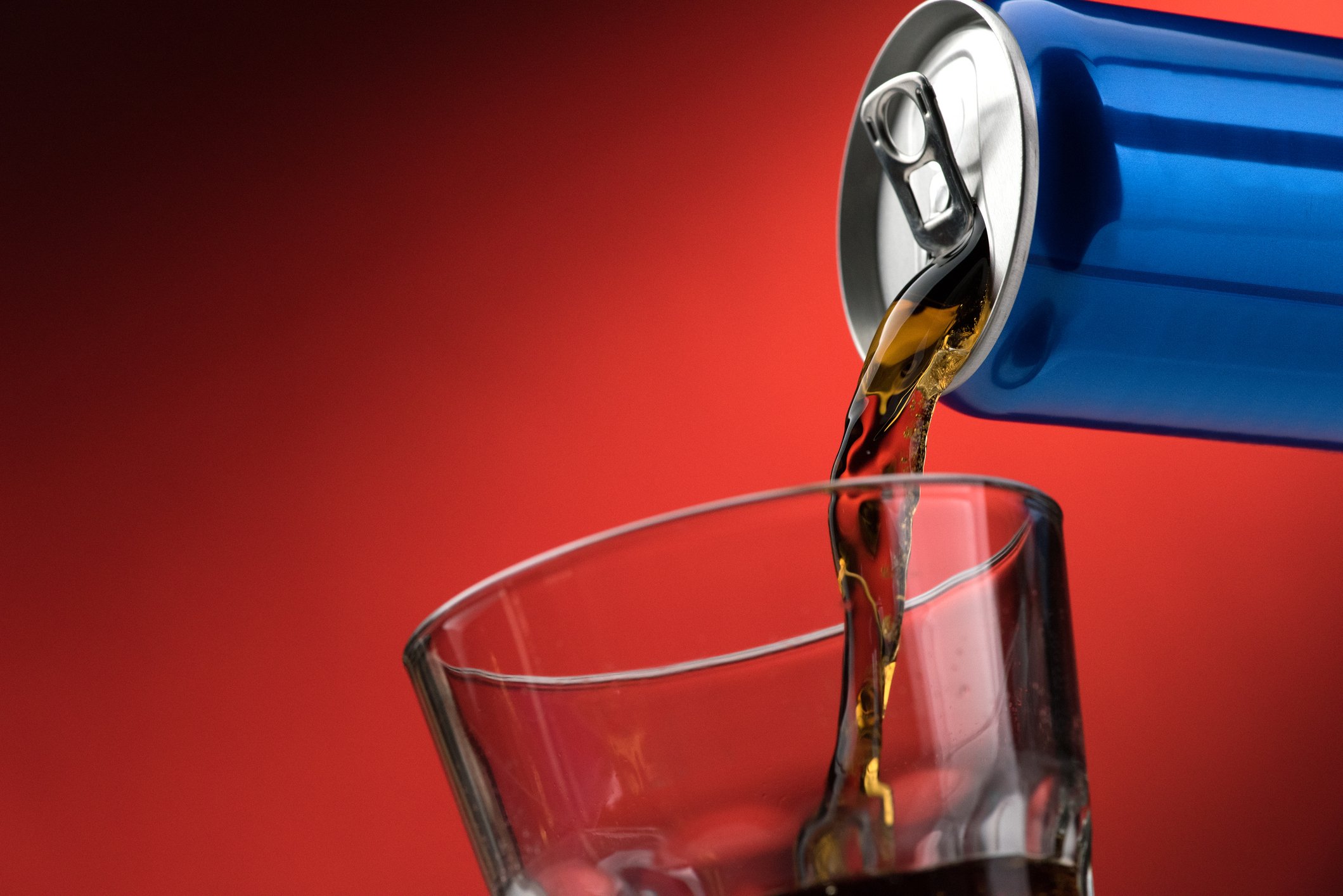 A soft drink being poured into a glass from a blue aluminum can, against a red background.