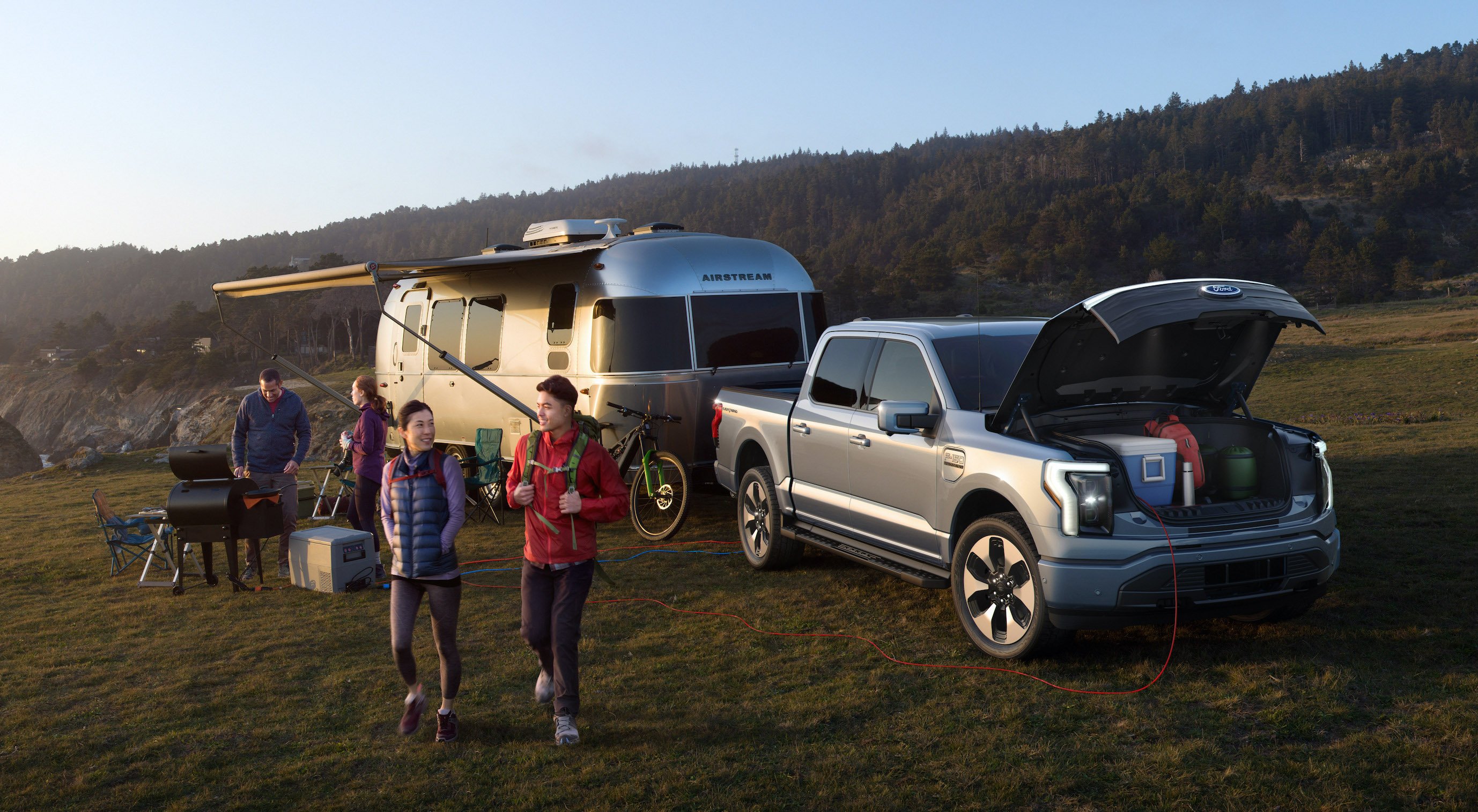 A silver Ford F-150 Lightning, an electric pickup truck, with an Airstream camping trailer at a campsite.