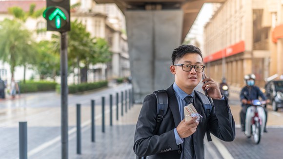 A young businessperson standing on a median, holding a sandwich and smartphone, and seemingly waiting for a ride. 