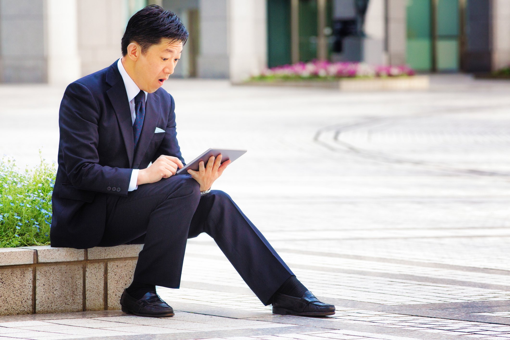 A person in a business suit looking at a tablet with surprise while sitting on a landscaping wall outside.
