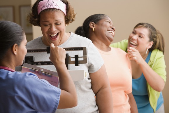 Three women smile as a medical worker weighs one of them on a scale.