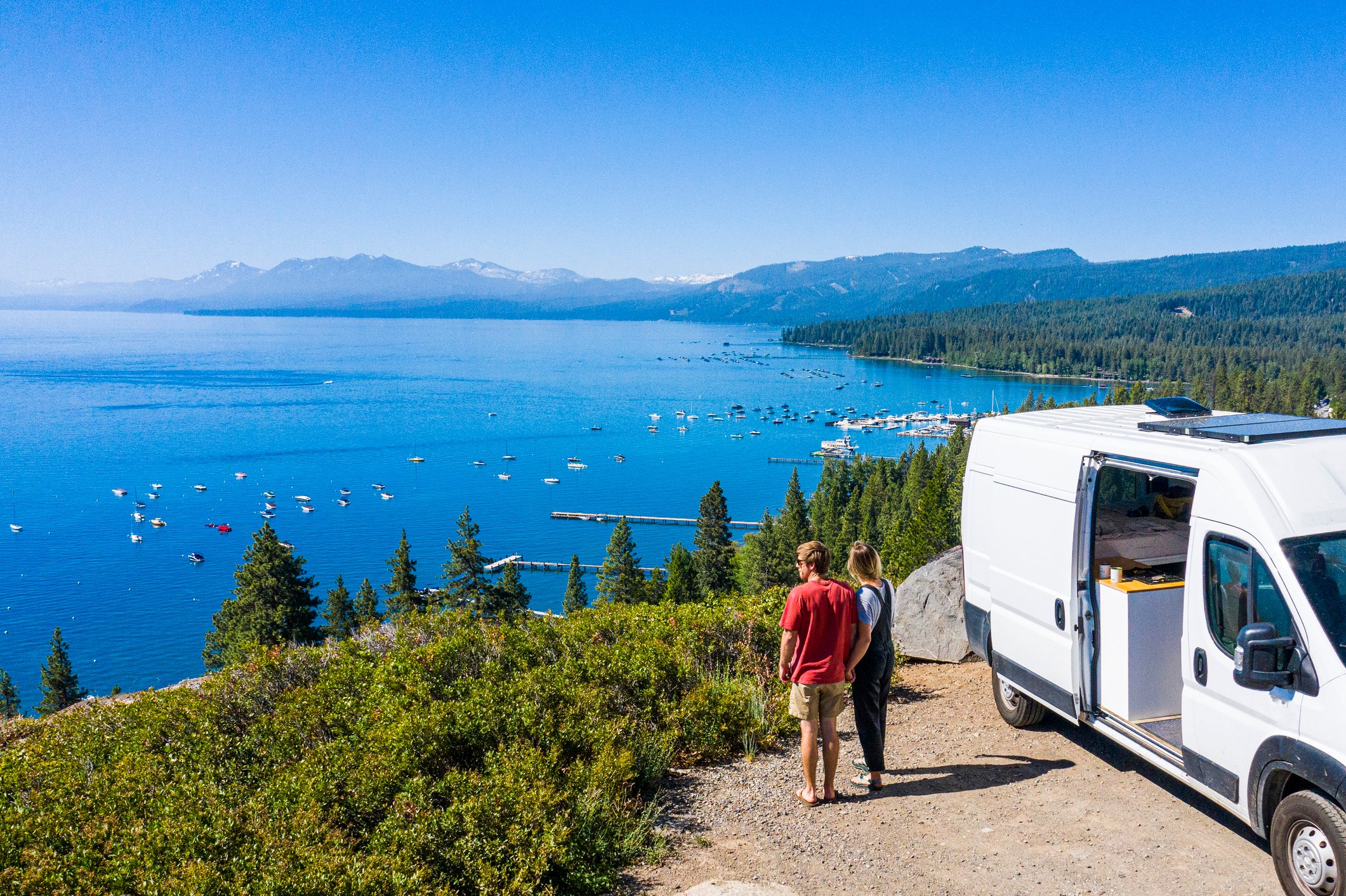 Two people standing next to RV on scenic overlook above Lake Tahoe. 