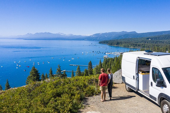Two people standing next to RV on scenic overlook above Lake Tahoe. 