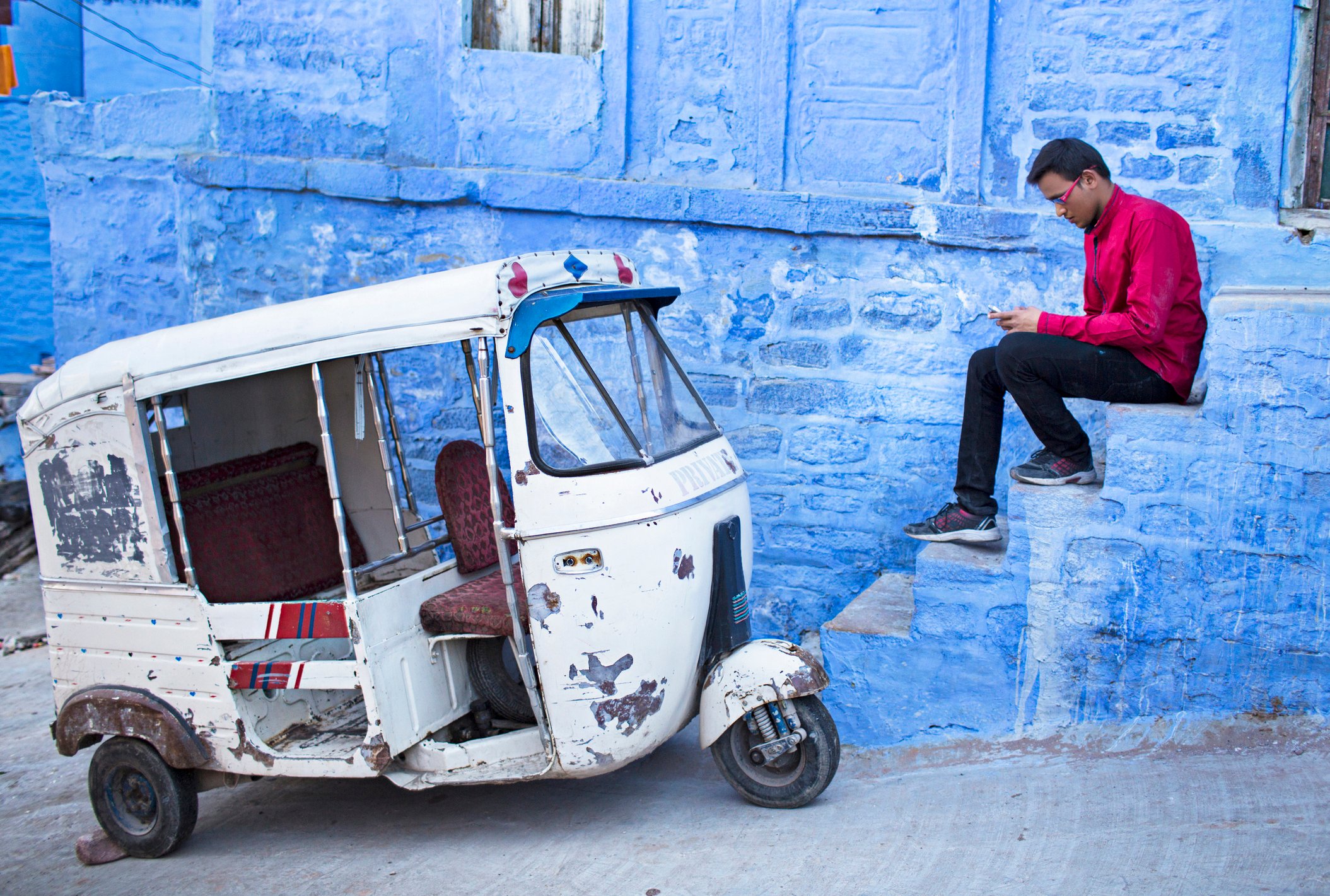 Person sitting on steps using a smartphone in front of a vehicle.