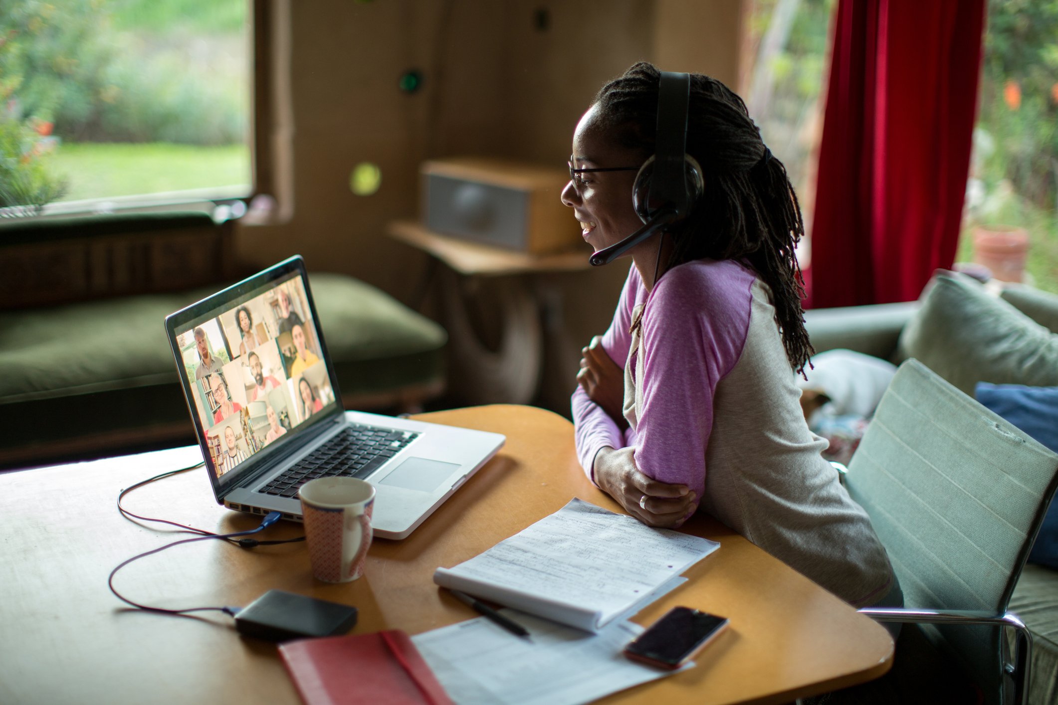 A student with a headset watches a videoconference on a laptop, with note-taking materials and a cup of coffee strewn around it.
