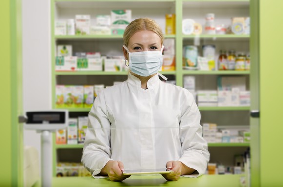Pharmacist wearing a face mask with medications on shelves in the background.