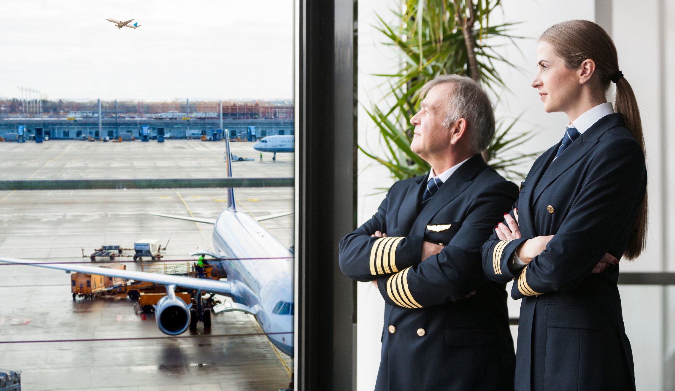 Two pilots looking out a window onto the tarmac, with an airplane in the background.