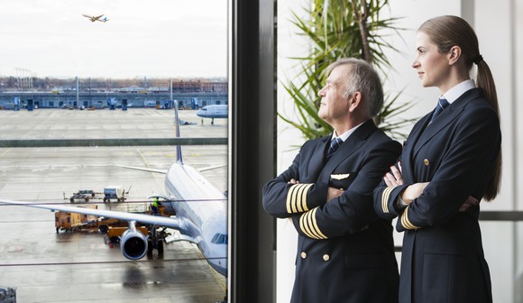 Two pilots looking out a window onto the tarmac, with an airplane in the background.