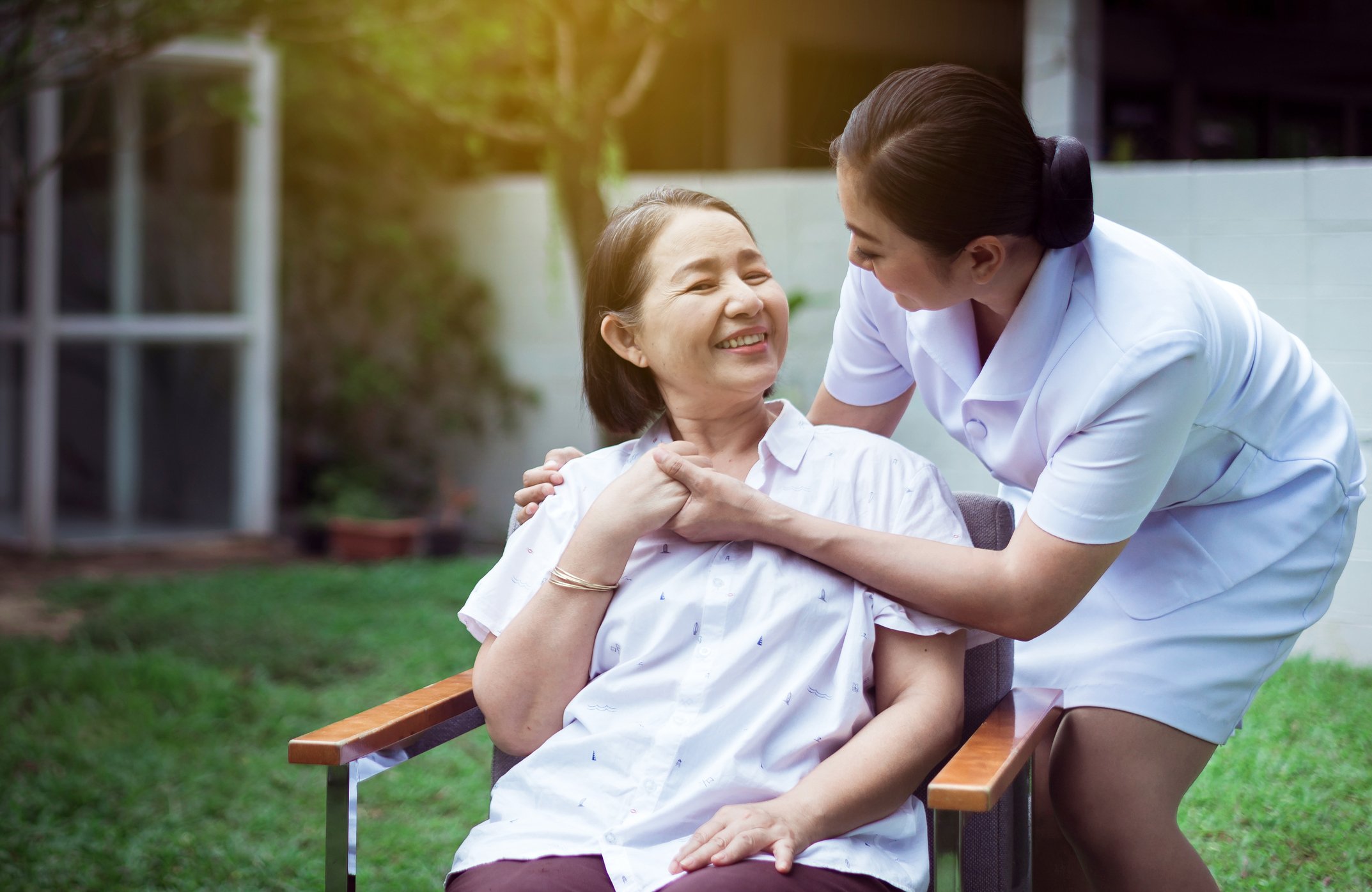A caretaker looks after a smiling patient sitting outside in a chair.
