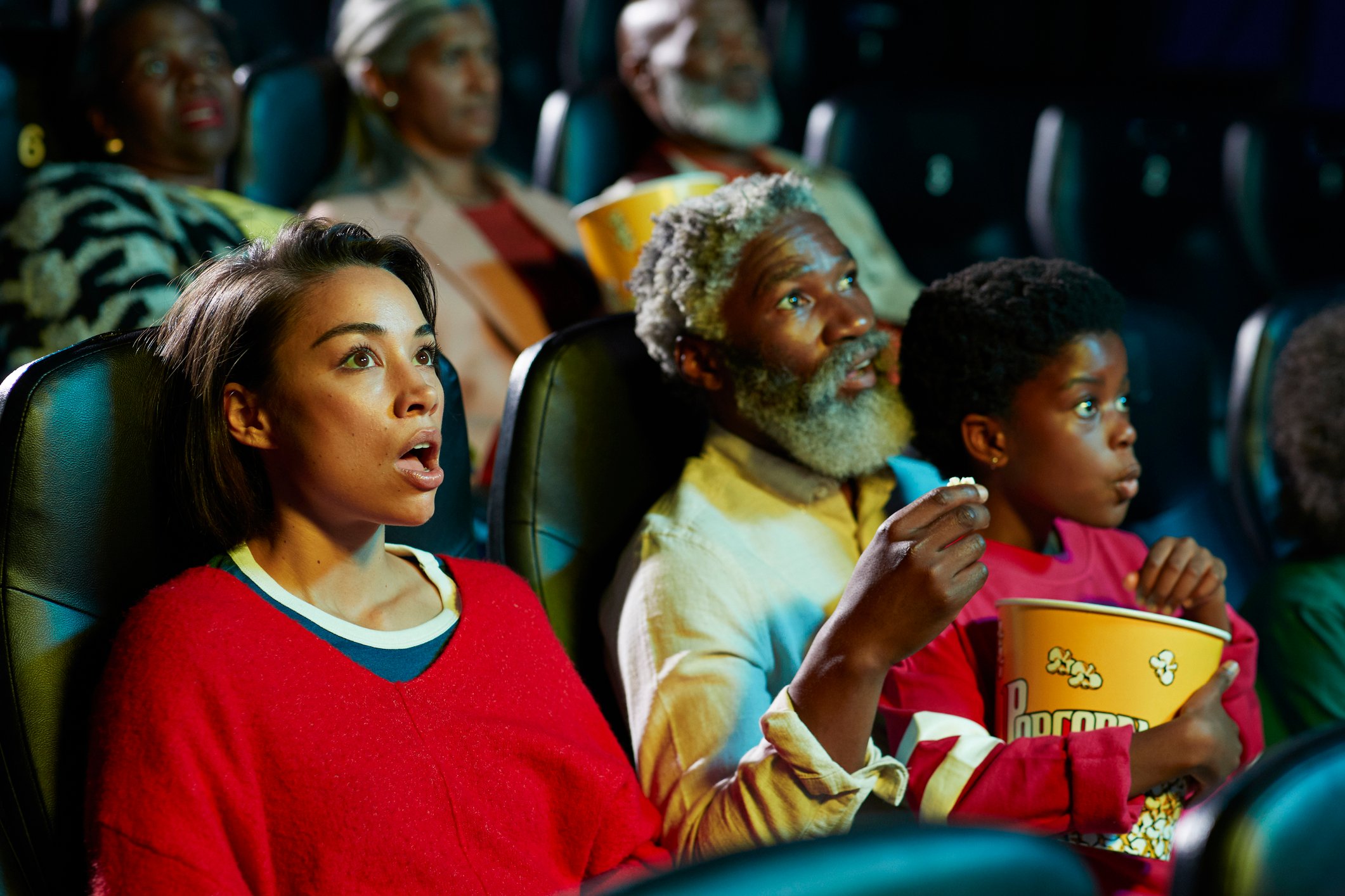 A parent, grandparent, and child holding a bucket of popcorn react with surprise while watching a movie.